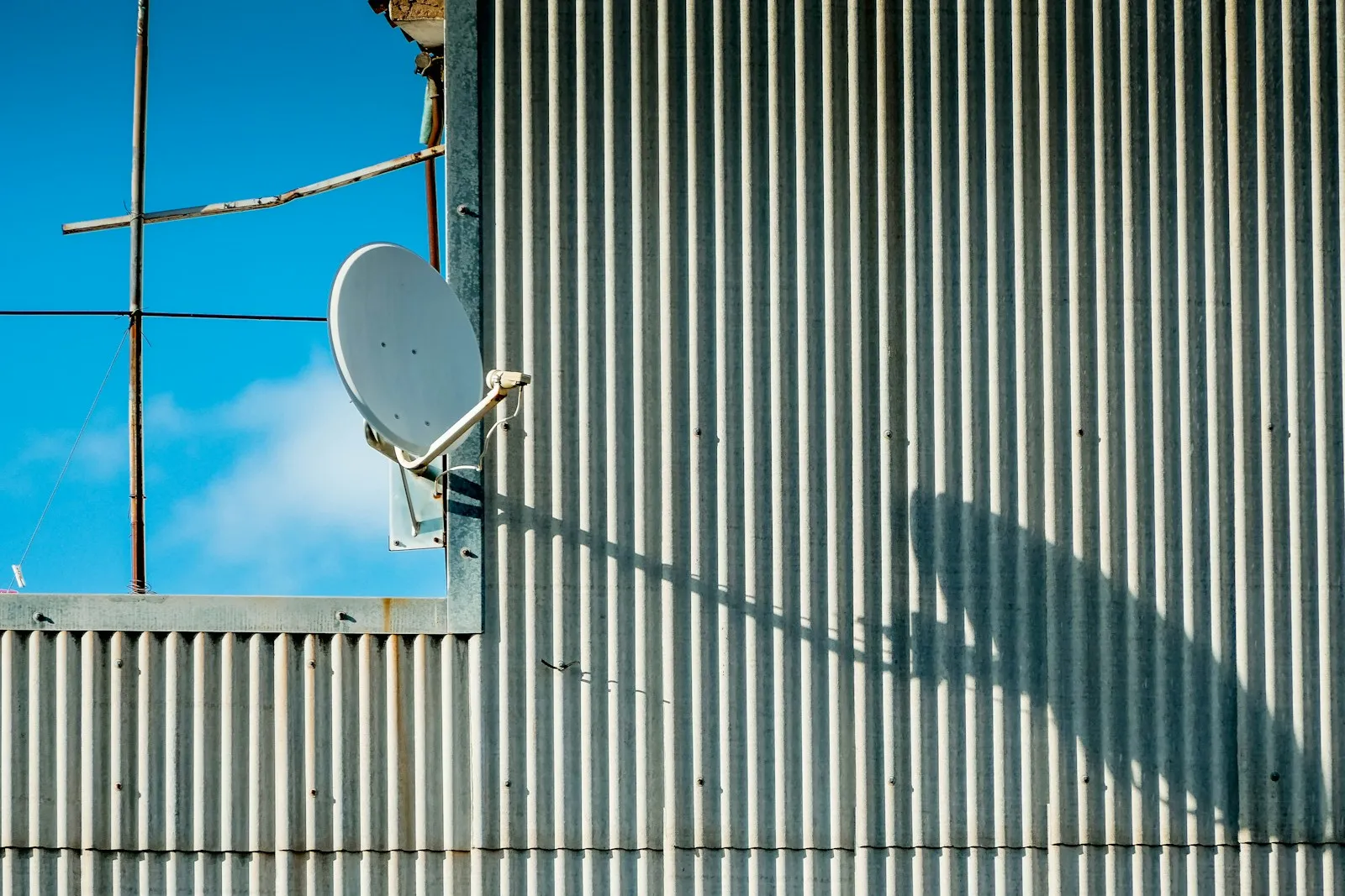 A satellite dish mounted on the corrugated wall of a building, against a blue sky.