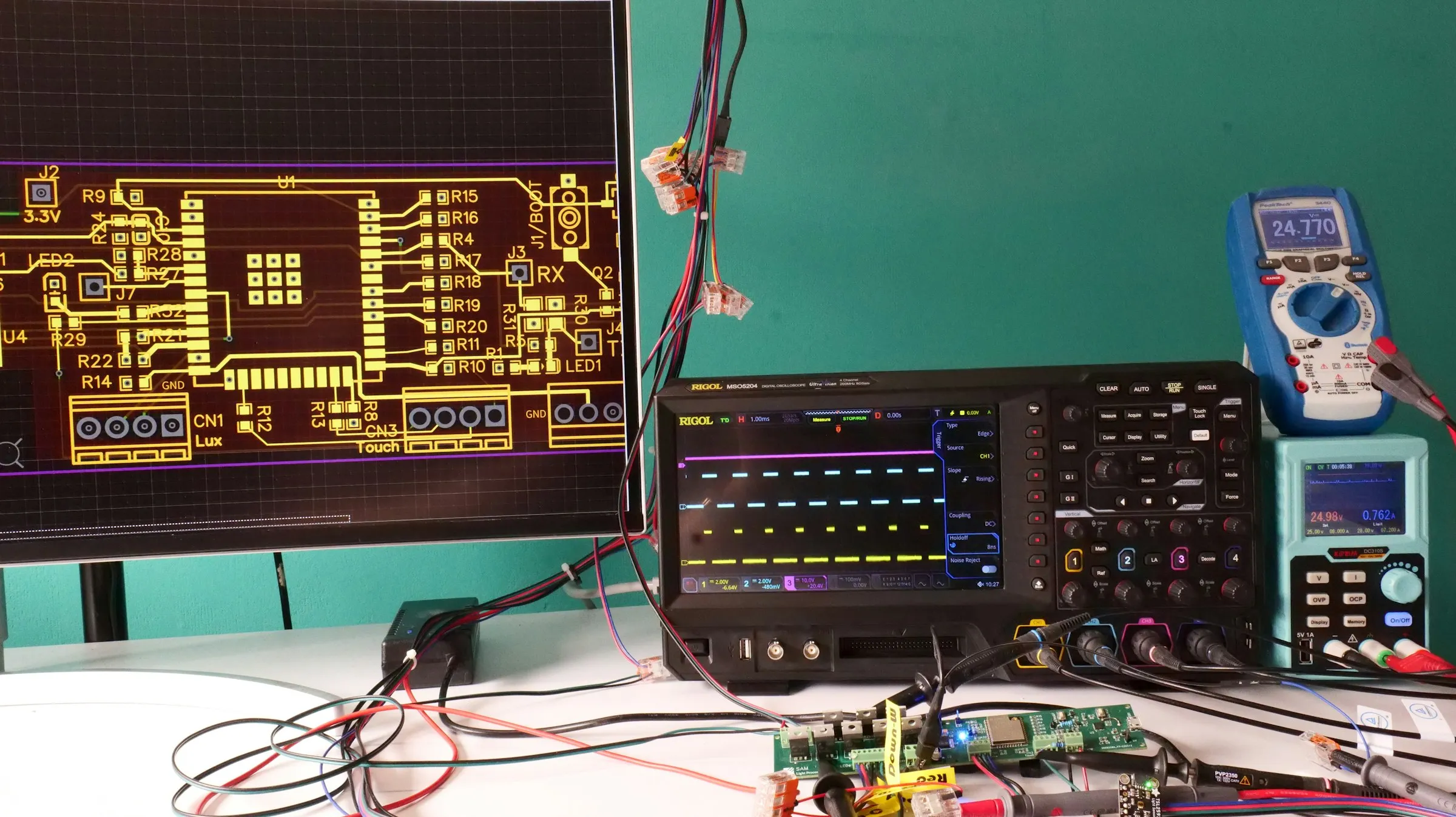 An electronics test bench: oscilloscope showing live waveforms, bench power supply with OVP and OCP controls, and a multimeter reading 24.770 V.